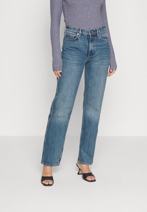 Kvinde iført højtaljede blå denimbukser, en figursyet grå langærmet top og sorte åbne hælede sandaler, stående mod en ensfarvet baggrund.