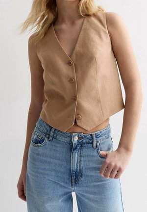 Femme portant un gilet beige sans manches boutonné et un jean bleu délavé taille haute, main reposant dans la poche du jean, sur fond uni.