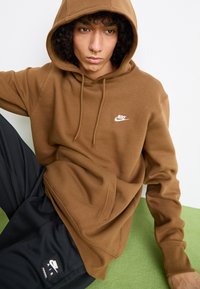 Brun Nike hettegenser med lommesfront og hvit logo. Kombinert med svarte bukser som har et Nike Air-merke. Myk tekstur.