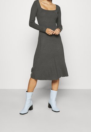 Femme portant une robe midi à manches longues gris foncé avec un décolleté carré et des bottes à talons bleu clair mi-mollet, debout sur un sol gris.