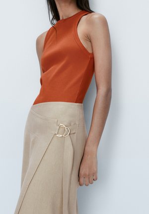 Persoon die een mouwloos topje in verbrande oranje kleur en een beige wikkelrok met gouden gesp draagt, staand tegen een lichtgrijze achtergrond.