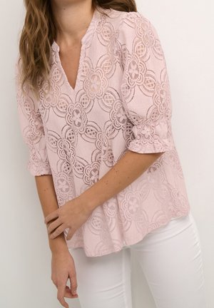 Donna che indossa una camicetta in pizzo rosa chiaro con motivi floreali e maniche a tre quarti abbinata a pantaloni bianchi su uno sfondo semplice.