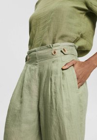 Mujer vistiendo pantalones de lino de cintura alta en verde claro con bolsillos laterales y botones, con la mano descansando en el bolsillo, combinados con una blusa verde a juego.