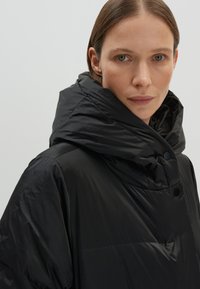 Zwarte gewatteerde jas met een hoge kraag en grote capuchon. Beschikt over een gladde, glanzende stof, drukknoopsluiting en een minimalistisch ontwerp.