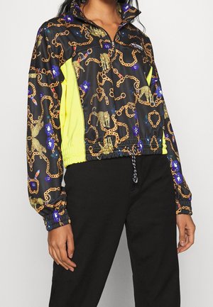 Chaqueta negra de manga larga con cadena dorada y estampado de leopardo, flores azules, paneles laterales amarillos, cierre de cremallera en el cuello, combinada con pantalones negros.