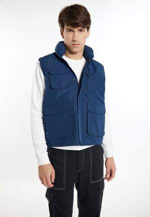 Navyblauwe gewatteerde vest met hoge kraag, meerdere frontzakken, ritsluiting en elastische zoom, gedragen over een witte blouse met lange mouwen.