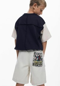 Jongen in een marineblauwe shirt over een beige shirt met korte mouwen en beige shorts met een kleurrijk Amazing Spider-Man stripboek pocketontwerp.