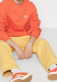 Pomarańczowa bluza z logo, żółte spodnie z kordurowej tkaniny i czerwone trampki z białymi paskami. Materiał wydaje się miękki i teksturowany.