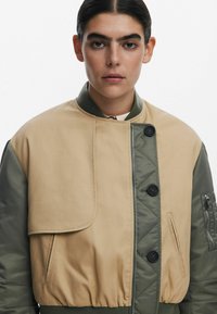 Beige en groene bomberjack met een ritssluiting aan de voorkant, twee grote zakken, geribde boorden en een flapdetail op de borst. Gewatteerde stof.