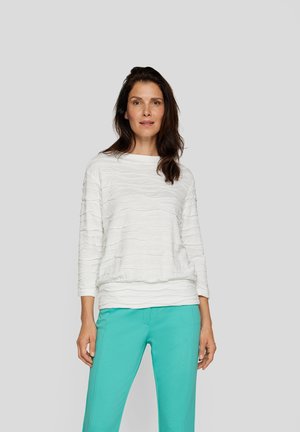 Weißes strukturiertes Langarmoberteil mit einem wellenförmigen Muster, kombiniert mit türkisfarbenen Hosen. Das Oberteil hat einen runden Halsausschnitt und einen gerafften Saum.