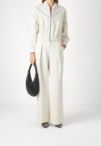 Ljuskräm bomberjacka med perforerat mönster, matchad med vita vida byxor, håller en svart läderhandväska och bär spetsiga metalliska skor.