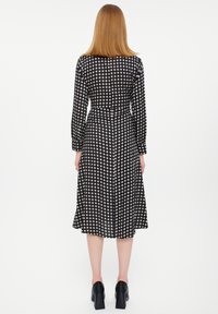Zwarte jurk met lange mouwen, voorzien van een geometrisch wit patroon. De stof heeft een gladde textuur, met een getailleerde taille en een uitlopende rok.