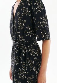 Gros plan sur une personne portant une robe noire à fleurs avec des manches courtes et une ceinture nouée à la taille, sur fond blanc.