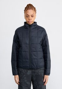 Veste matelassée noire avec un col haut, dotée d'un design matelassé, fermeture éclair frontale intégrale et coupe légèrement ample. Convient pour les superpositions.