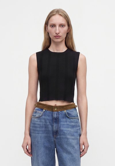 Jeune femme aux cheveux blonds raides portant un crop top noir sans manches et un jean bleu taille haute avec une ceinture marron.