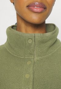Olivgrüner Fleece-Pullover mit Stehkragen, der eine strukturierte Oberfläche und fünf tonale Knöpfe an der Vorderseite hat.