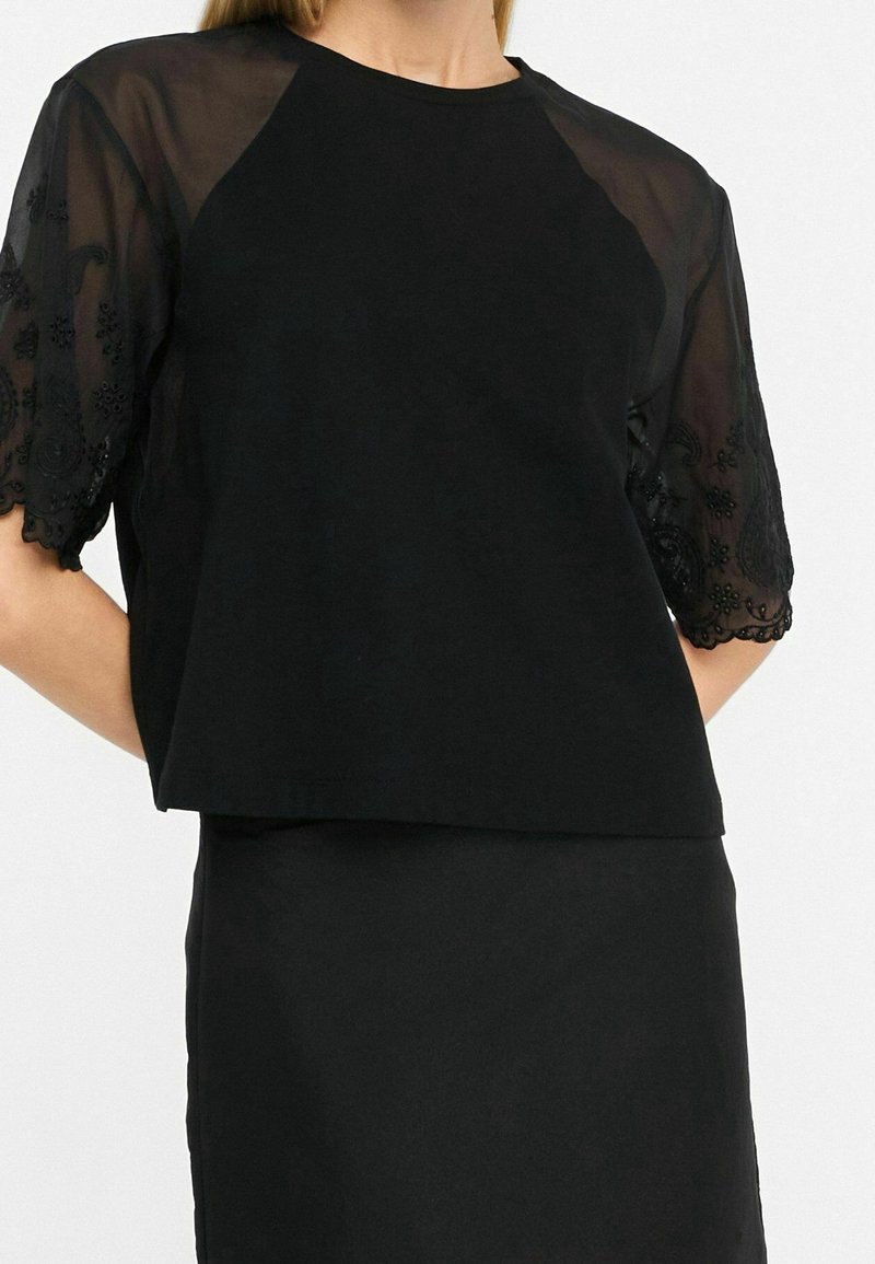 Femme portant un haut noir avec des manches courtes transparentes brodées et une jupe noire assortie, debout devant un fond blanc.