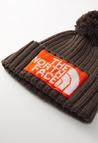 Brun strikket lue med en pom-pom på toppen. Har en livlig oransje lapp med "THE NORTH FACE" i hvite bokstaver. Strukturert ribbestrikket design.