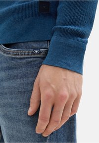 Suéter de punto azul con un patrón texturizado y puños acanalados, combinado con jeans de un azul claro que muestran un pequeño logo en el bolsillo trasero.