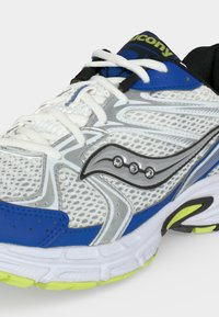 Κοντινό πλάνο ενός λευκού και μπλε παπουτσιού τρεξίματος Saucony με δίχτυ, λευκά κορδόνια και κίτρινο-πράσινη λεπτομέρεια στη σόλα.