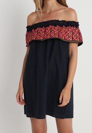 Donna che indossa un abito nero con spalle scoperte e una ampia balza ricamata con motivi rosa e arancioni sullo scollo.