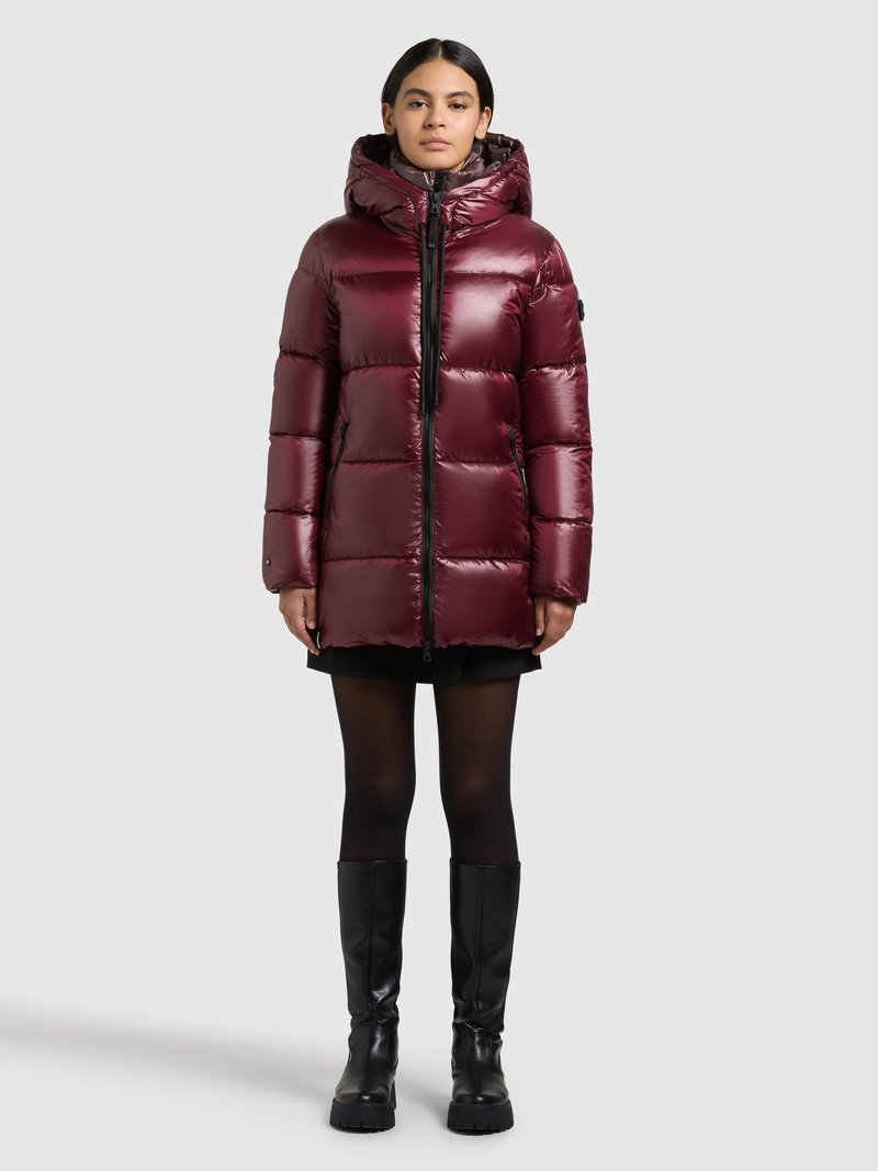 Viininpunainen toppatakki kiiltävällä pinnalla, hupulla, etusvetoketjulla ja tikatulla kuviolla. Yhdistetty mustaan hameeseen ja polviin ulottuviin mustiin saappaisiin.