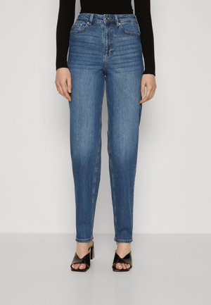 Kobieta w niebieskich dżinsach z wysokim stanem i czarnych sandałach na obcasie z odkrytymi palcami, stoi na jednolitym jasnym tle.