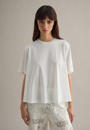 Donna con capelli castani lisci, che indossa una maglietta bianca a maniche corte e pantaloni bianchi con motivo a pizzo, in piedi davanti a uno sfondo semplice.