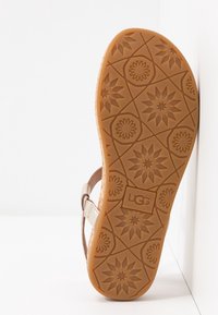 Ruskea kumipohja, jossa on rakennekuviointi kukista ja kohokuvioitu 'UGG' -logo. Kevyet vaaleanruskeat hihnat, joissa on metalliset yksityiskohdat.