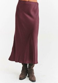 Dlouhá hladká burgundská sukně v kombinaci s hnědými semišovými botami na střední lýtka se sponami a nízkými podpatky na jednobarevném bílém pozadí.