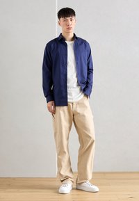 Cămașă bleumarin cu nasturi peste un tricou alb, asortată cu pantaloni cargo bej și adidași albi. Cămașa are mâneci lungi și o croială lejeră.