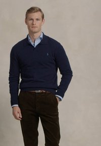 Pulover bleumarin cu guler, purtat peste o cămașă de culoare albastru deschis. Pantaloni din catifea de culoare maro și o curea maro. Design simplu, cu detalii de branding minime.