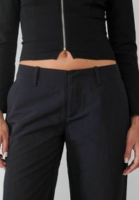 Zwarte cropped zip-up top met een split aan de onderkant boven de taille, gecombineerd met hoge zwarte broeken met strakke, gestructureerde lijnen en zakken.