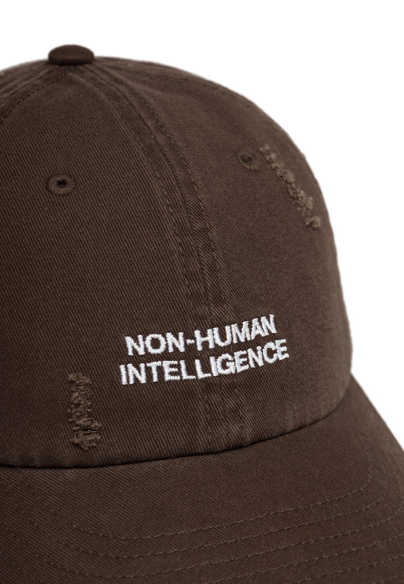 Cappello marrone in cotone, con testo ricamato bianco "NON-HUMAN INTELLIGENCE" e dettagli usurati. Visiera curvata e chiusura regolabile.