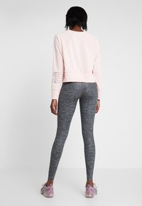 Felpa cropped rosa con maniche raglan e lettering "NIKE" sul braccio sinistro, abbinata a leggings grigi scuri con texture e sneakers rosa chiaro.