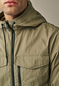 Veste à capuche vert olive en tissu texturé, dotée d'un zip sur le devant, d'accents noirs et de deux poches poitrine.