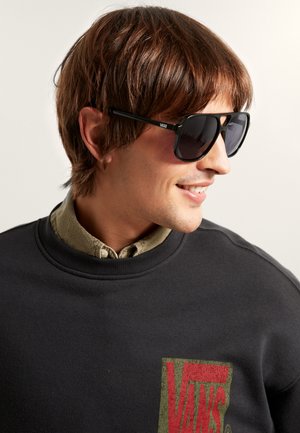 Hombre con cabello castaño que lleva gafas de sol Vans negras, camisa de botones color oliva y sudadera negra con el logo rojo de Vans, sonriendo y mirando a la derecha.