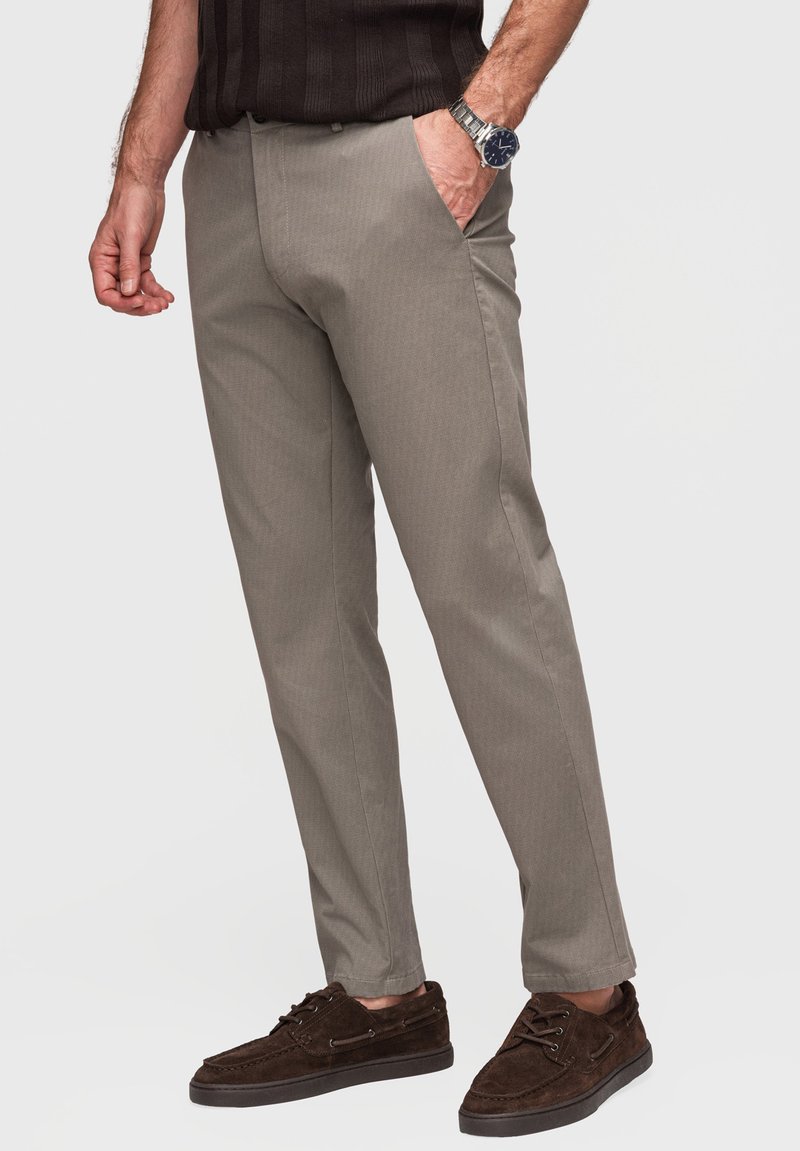 Homme portant un pantalon beige droit, des chaussures décontractées marron foncé, une chemise sombre et une montre-bracelet argentée, debout avec une main dans la poche.