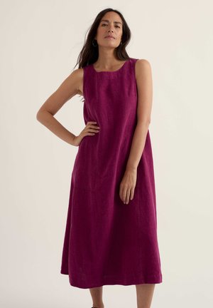 Vrouw in mouwloze, diep magenta midi-jurk staand met één hand op de heup tegen een effen lichte achtergrond.