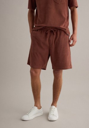 Hombre con pantalones cortos de rizo marrón con cordón y parte superior a juego, combinados con zapatillas blancas bajas sobre fondo neutro.