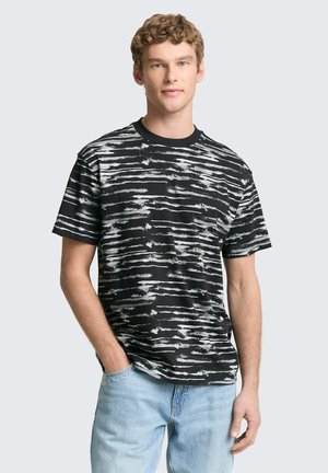Junger Mann mit lockigem Haar, trägt ein schwarz-weiß gestreiftes kurzärmeliges T-Shirt und hellblaue Jeans, steht mit einer Hand in der Tasche.