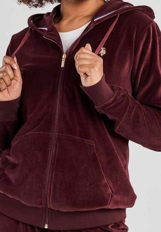 Osoba v zapínací bordeaux sametové mikině s předními kapsami a ribbovanými manžetami, s rukama zvednutýma u hrudníku, ukazující detaily oděvu.