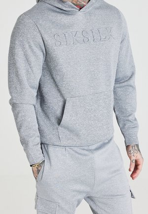 Mężczyzna w jasnoszarej bluzie z kapturem i dopasowanych spodniach dresowych z wytłoczonym na piersi "SIKSILK" oraz widocznymi tatuażami na dłoniach i bransoletką.