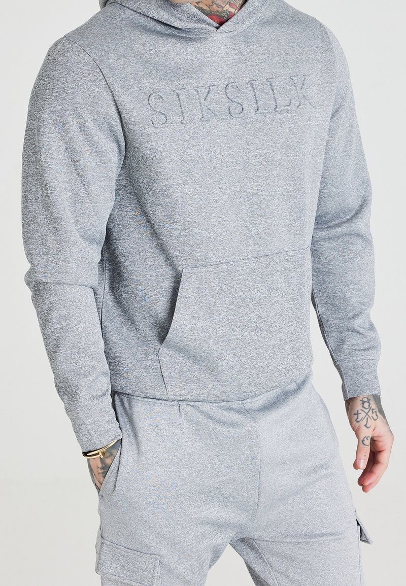 Homme portant un sweat à capuche gris clair et un pantalon de jogging assorti avec "SIKSILK" embossé sur la poitrine et des tatouages visibles sur les mains ainsi qu'un bracelet.