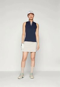 Tamsiai mėlynas be rankovių polo marškinėliai su užtrauktuku kaklo srityje, derinami su šviesiai smėlio spalvos golfo sijonu, balta kepure, pirštine ir šviesios spalvos golfo bateliais.