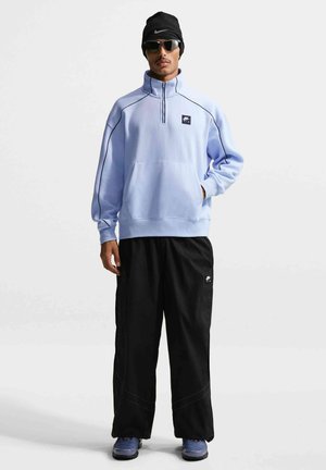 Mees, kes kannab helehalli poollukuga dressipluusi, mustaid laiaid pükse, musta talvemütsi, päikeseprille ja siniseid tossusid, seisab ühevärvilise valge tausta ees.