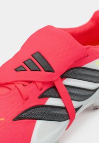 Λαμπερό ροζ ποδοσφαιρικό παπούτσι με μαύρες λεπτομέρειες, που διαθέτει υφή στο πάνω μέρος, κομψό σχέδιο και διακριτικό λογότυπο με τρεις ραβδώσεις.