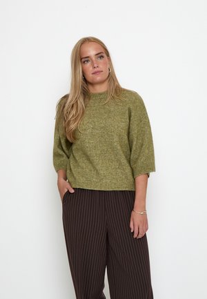 Jonge vrouw met lang blond haar, gekleed in een groene gebreide trui en donkere broek met krijtstrepen, staand met één hand in de zak.