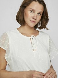Blusa bianca con motivo a pois testurizzato, maniche corte a campana e scollo a goccia legato con un nastro di tessuto.