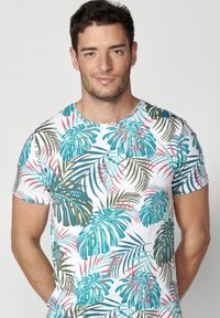 Camiseta de manga corta con un vibrante patrón de hojas en turquesa, verde y rosa sobre un fondo blanco, hecha de una tela ligera.